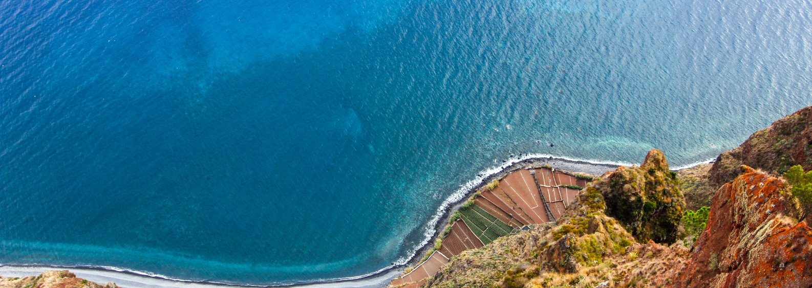 Aerial view of deep blue sea curving along a narrow shoreline with terraced coastal fields and rugged reddish cliffs in foreground.
