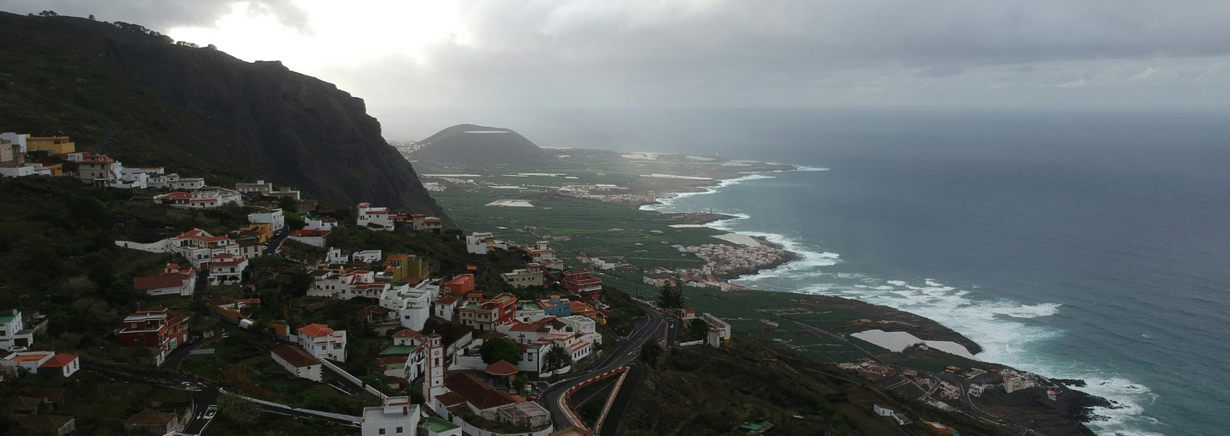 Hillside village of white and red-roofed houses clinging to cliffs above winding roads, green fields and a cloudy Atlantic coastline with surf.