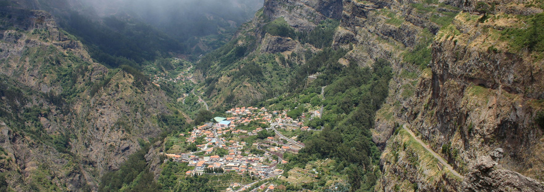 Aerial view of a red-roofed village tucked into a steep, green mountain valley with winding roads and rocky cliffs.