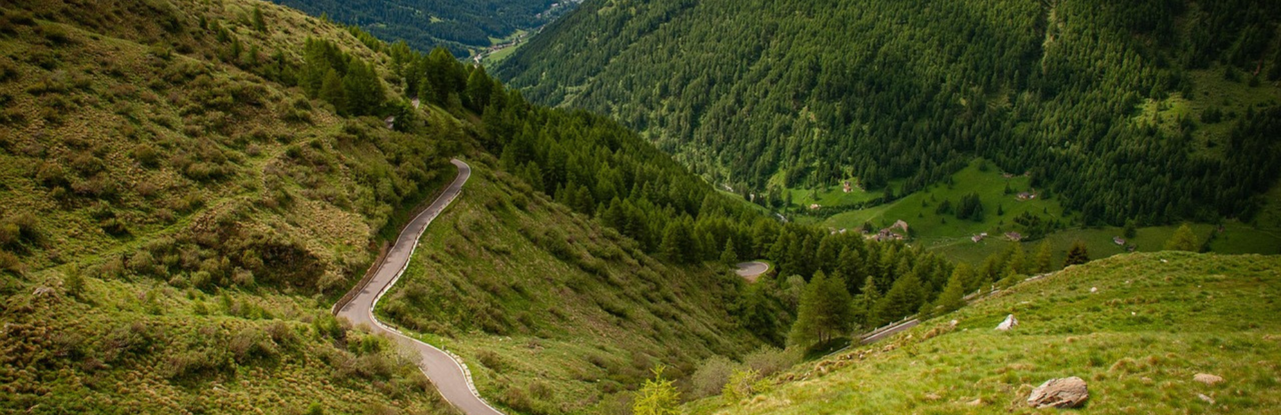 Winding mountain road with hairpin turns descending through green hills and pine forests into a valley with scattered houses.