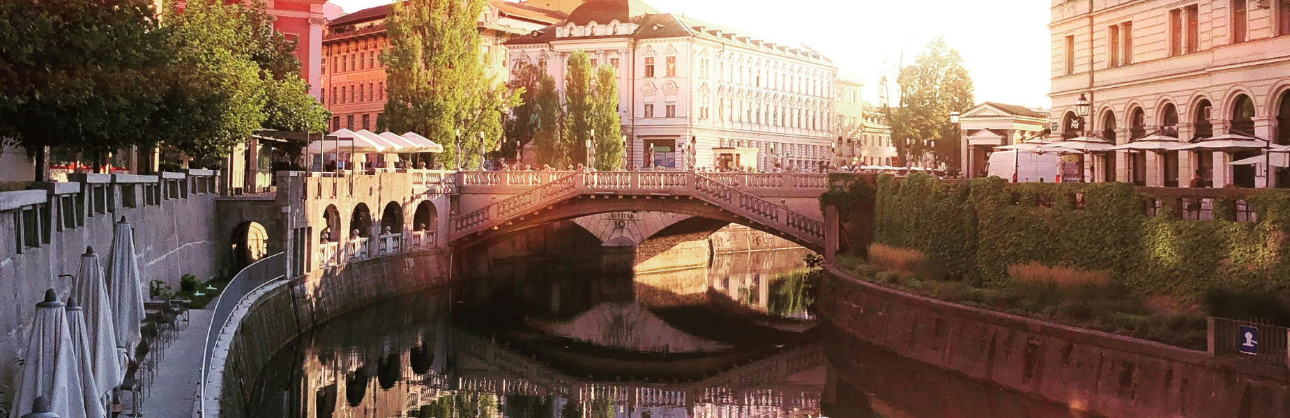 Sunlit canal with arched stone bridge, riverside cafés, trees and reflection of ornate buildings in calm water.