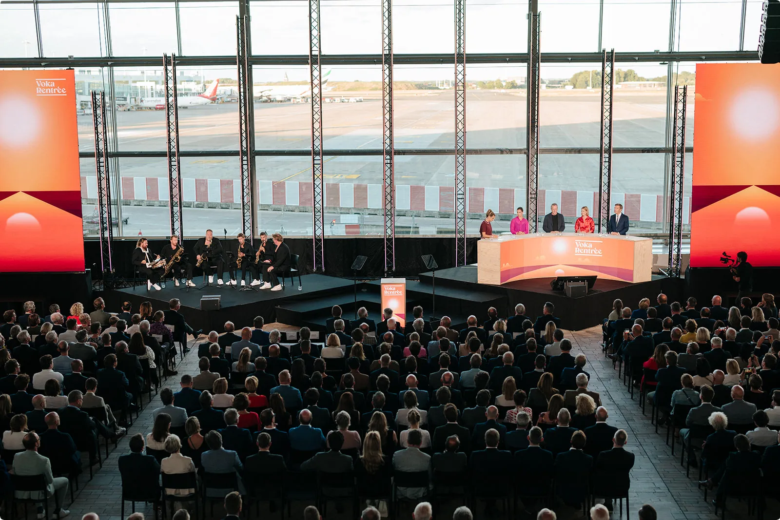 A large indoor event with a panel discussion and audience, set against a backdrop of airport windows showing aircraft and the runway.