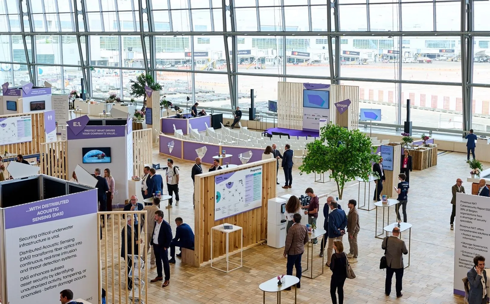 Large, open exhibition space with people networking around wooden booths and tables. Indoor trees and digital screens are placed throughout.