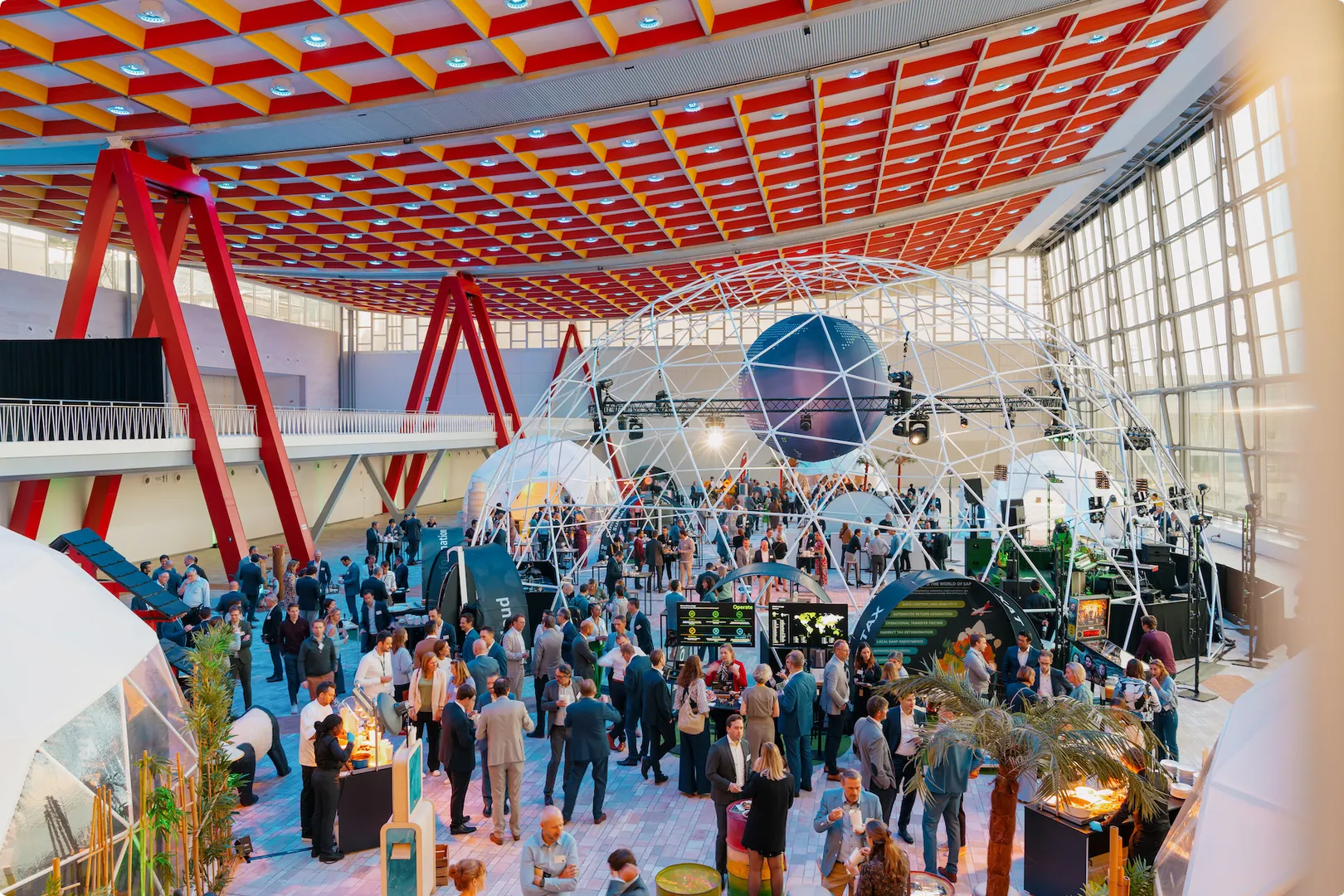 A large indoor event with people mingling under a geodesic dome, colorful ceiling, and red support beams in a modern, well-lit venue.