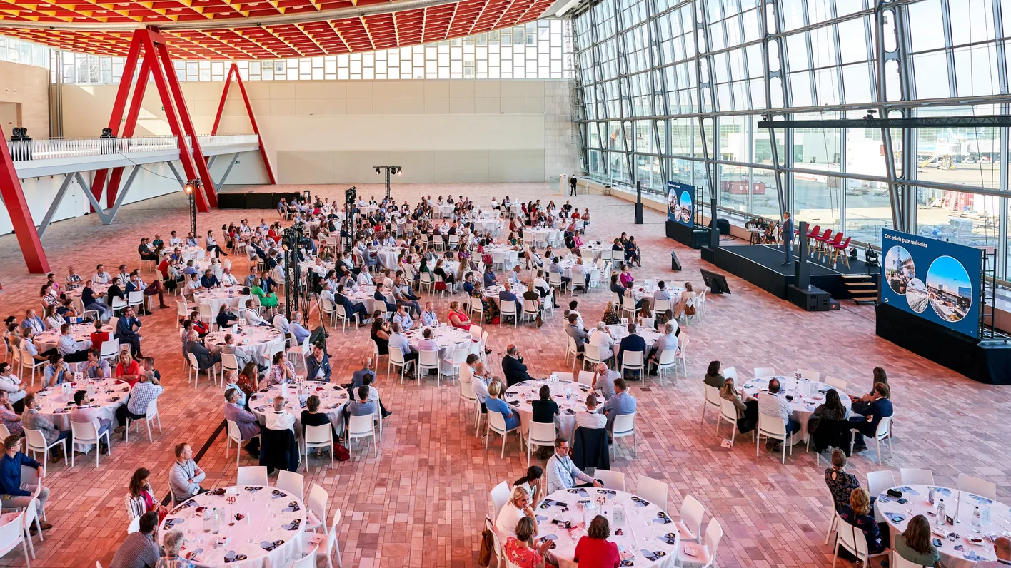 A large conference room with round tables set for a formal event. People are seated, and a speaker is on stage with presentation screens beside them.