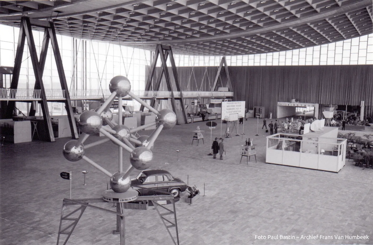 A black and white photo of a large exhibition hall with an Atomium model, a vintage car, and people walking around various displays.