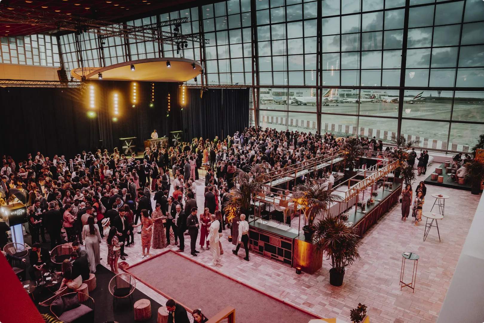 A large group of people attending a formal event in a spacious venue with large windows and a view of airplanes outside.