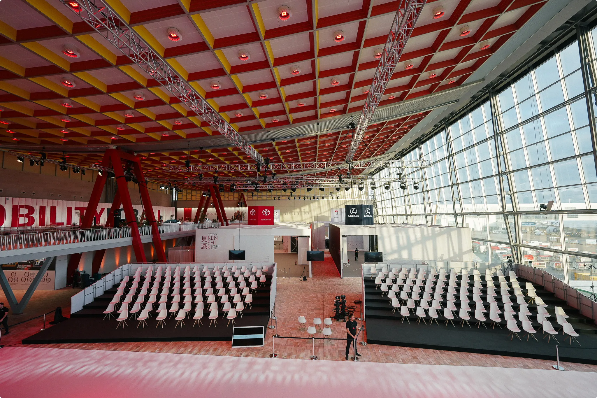 Spacious event hall with rows of white chairs, red and yellow ceiling, large windows, and branded banners on walls. Translucent flooring visible.