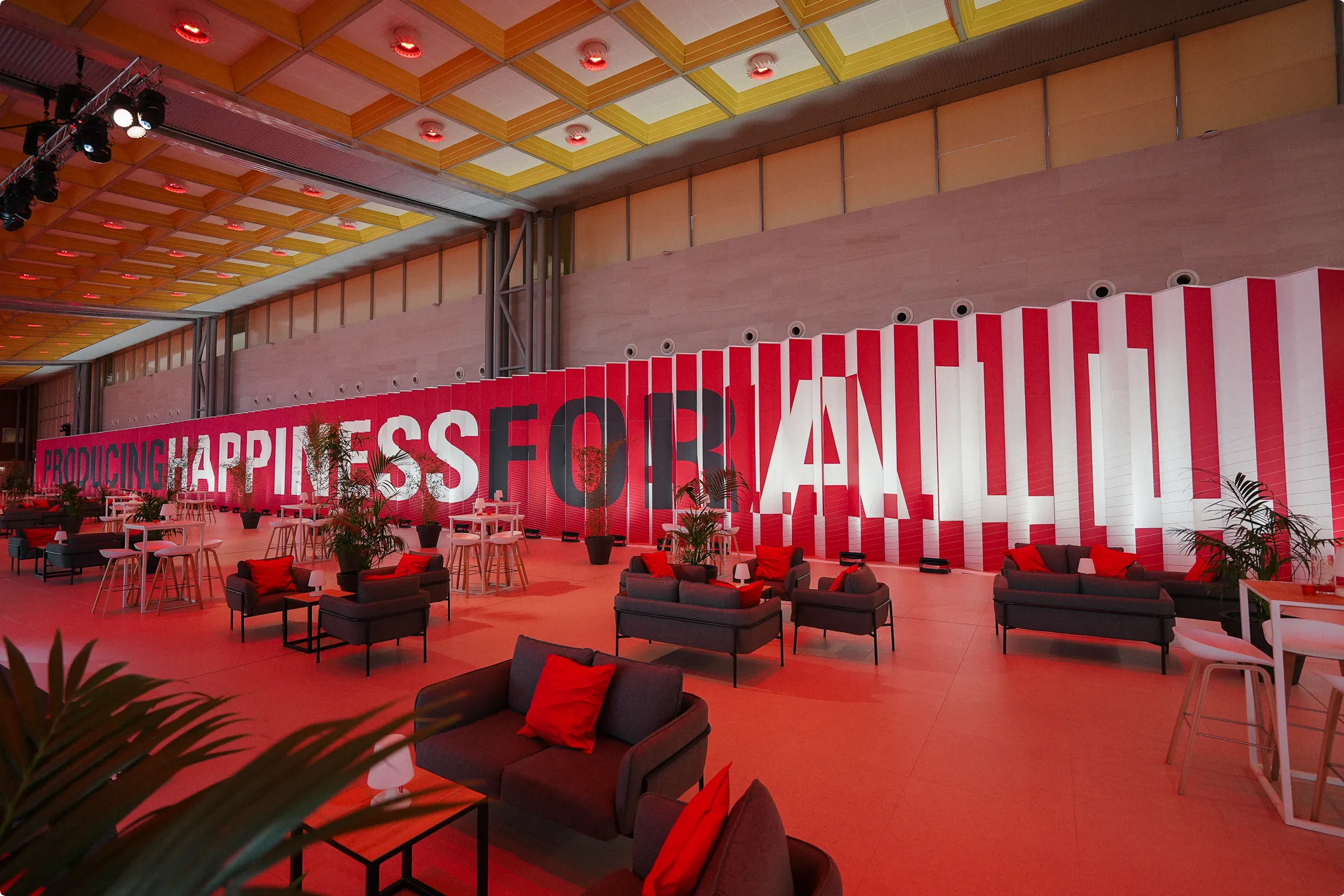 A modern, spacious lounge with red and gray sofas, plants, and a striped wall displaying the message "PRODUCING HAPPINESS FOR ALL" in bold letters.