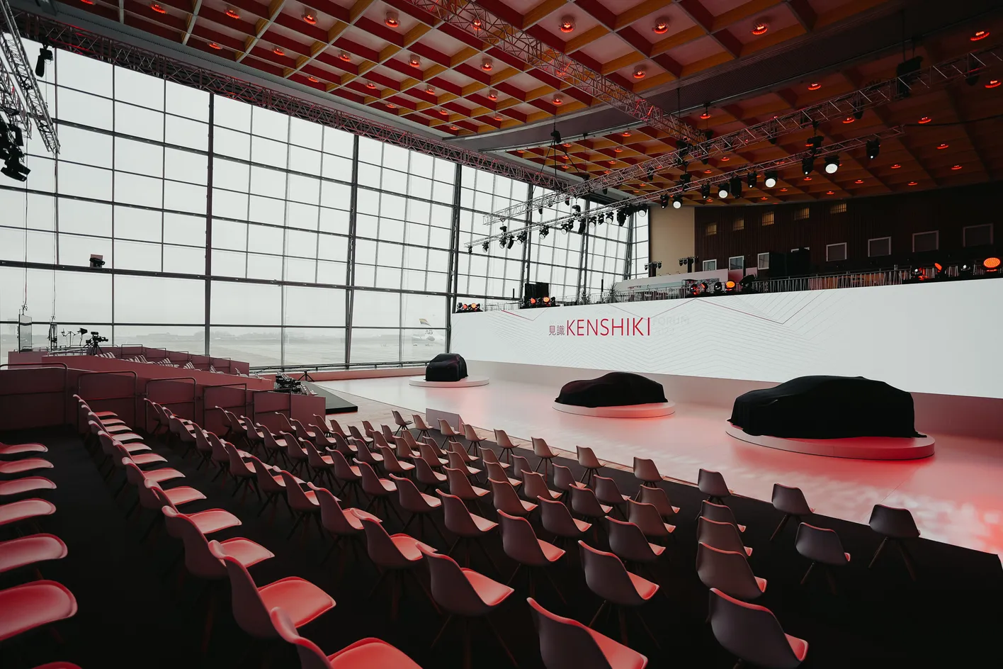 Empty event hall with rows of chairs facing a stage featuring two covered objects, large windows, and a sign reading "Kenshiki."