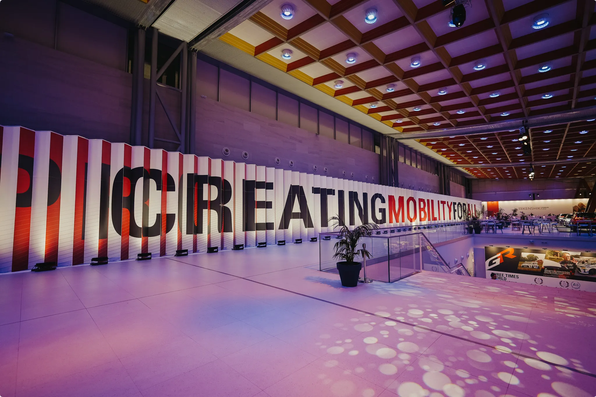 Large exhibition space with a ceiling featuring circular lights. A long wall displays "CREATING MOBILITY FOR ALL" with colorful lighting effects.