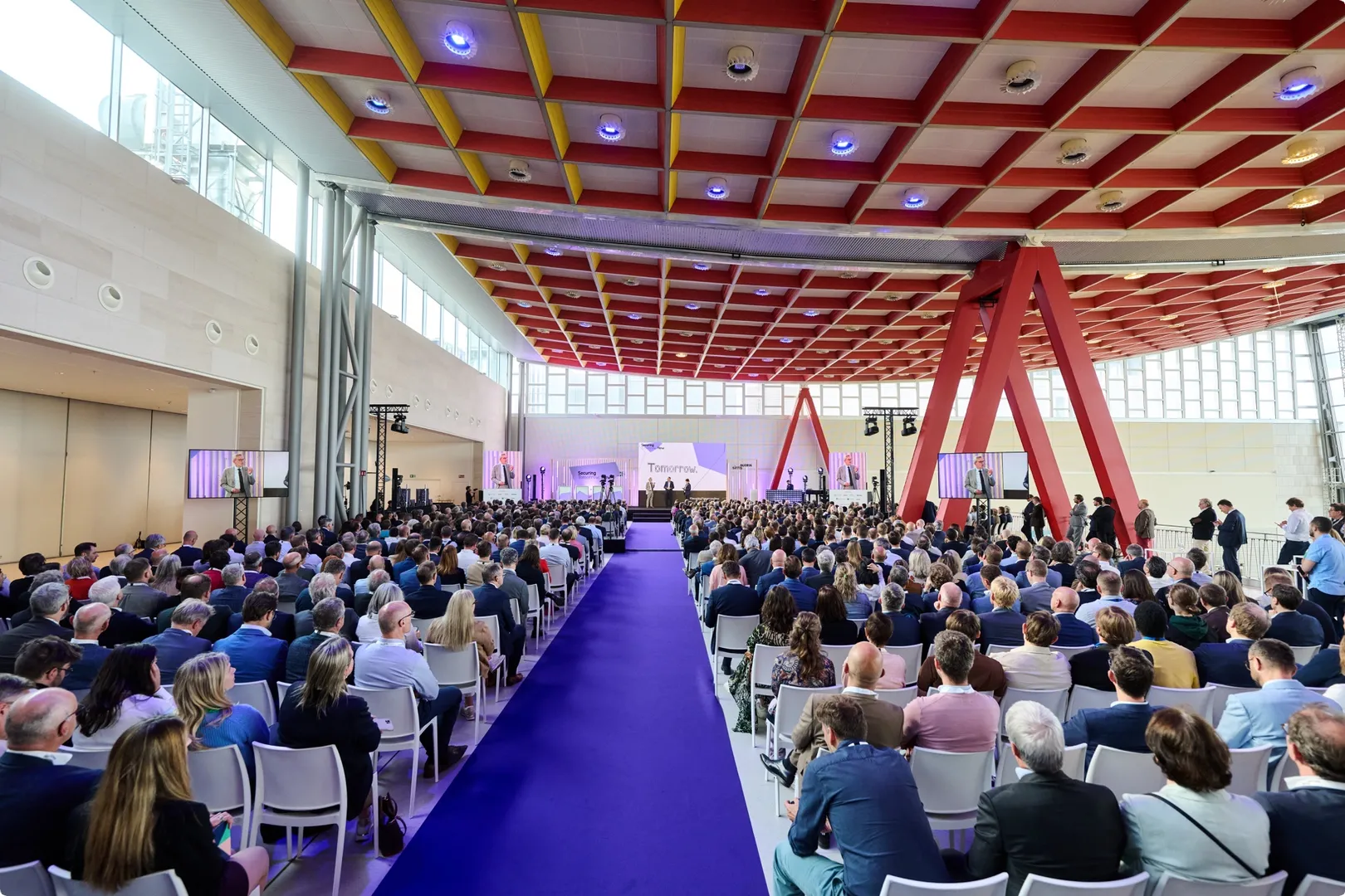 A large audience seated in a modern, architecturally unique conference hall with red beams and a purple carpet, listening to a presentation on stage.