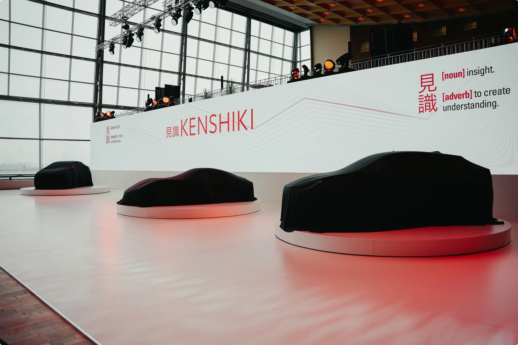 Three cars covered with black cloth on platforms in a showroom, with a large screen displaying "Kenshiki" and definitions for insight and understanding.