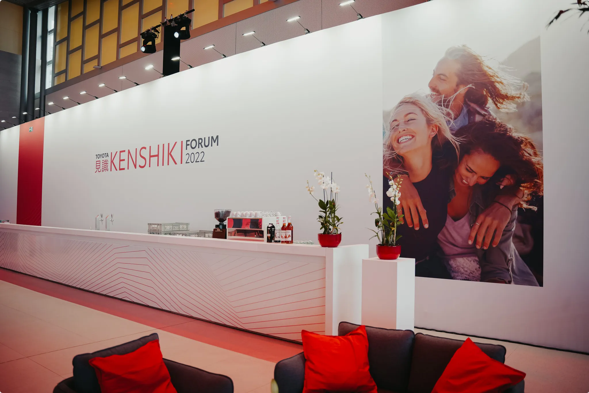Exhibition space with a large "Toyota Kenshiki Forum 2022" banner, featuring smiling people, modern decor, plants, and a long white counter.