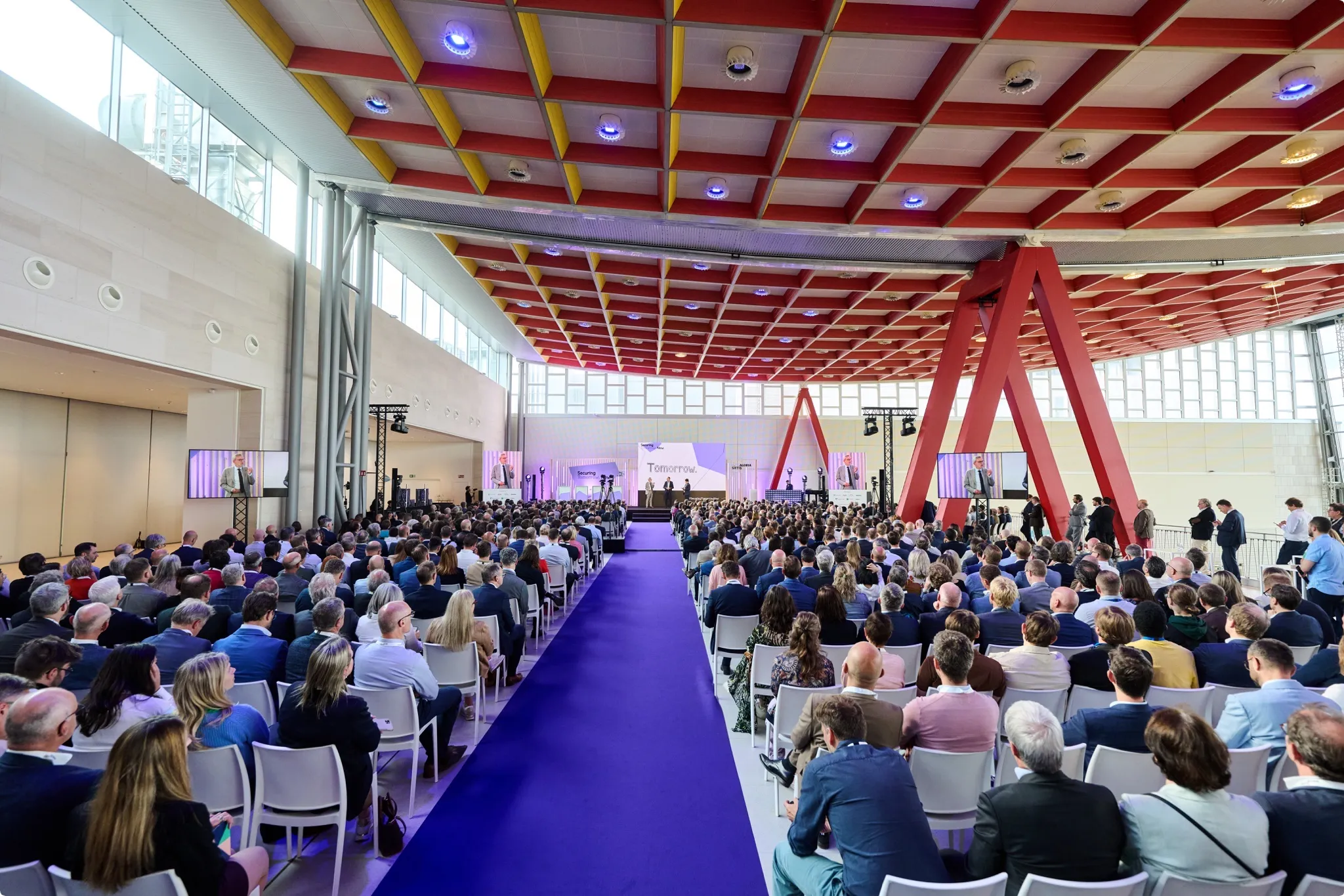 A large audience sits in a modern hall with a purple carpet, listening to a speaker on stage. The ceiling features a red and yellow grid design.