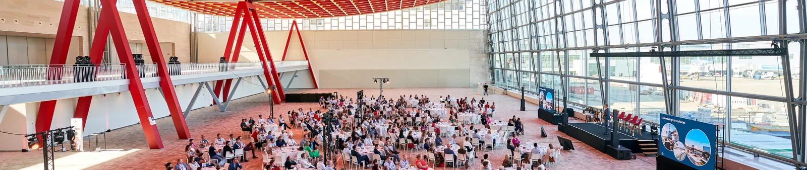 A large, modern conference hall with a glass wall hosts numerous people seated at tables, with red structural beams and a stage in view.