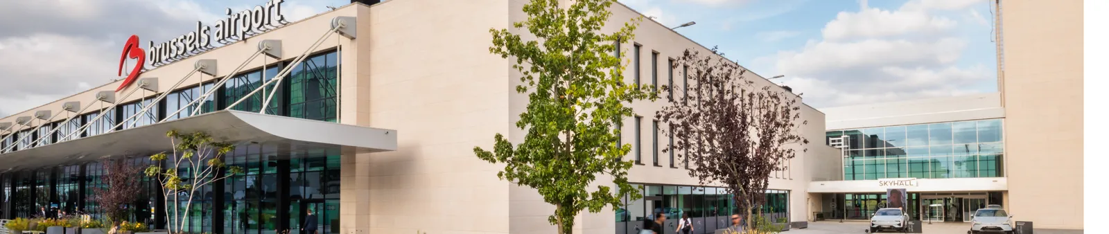 Exterior view of Brussels Airport with modern glass windows and beige facade, trees, and a clear blue sky with clouds.