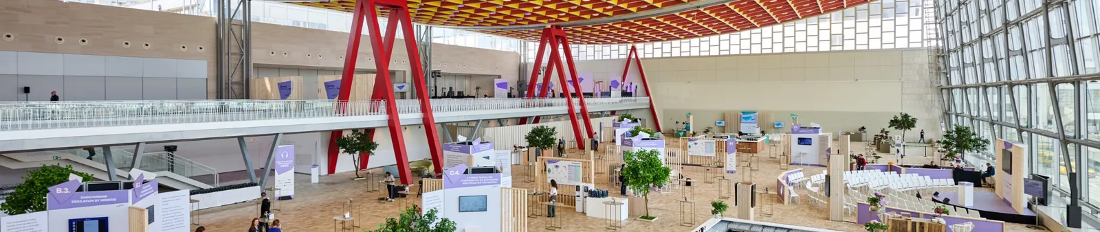 Spacious convention hall with red beams, multiple exhibition booths, potted trees, and rows of chairs under a patterned ceiling.