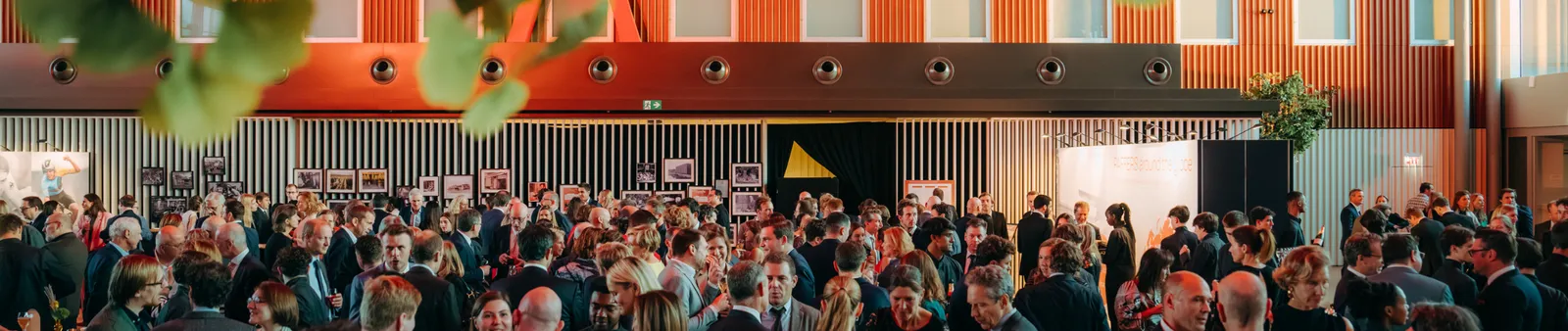 A large crowd of people socializing at an indoor event with framed photos on the walls, under warm lighting, with green leaves in the foreground.