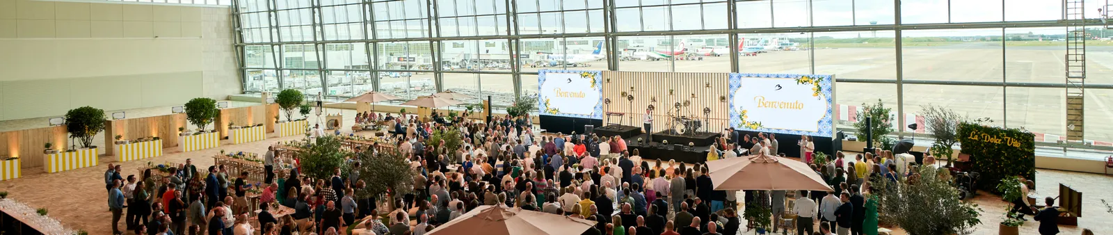 A large indoor gathering with people mingling near a stage, under a glass wall overlooking an airport runway. Screens display "Benvenuto."