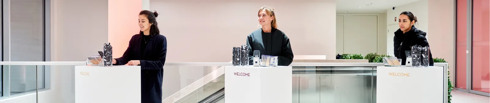 Three women stand behind welcome counters in a sleek, modern interior, each with water bottles and promotional materials on display.