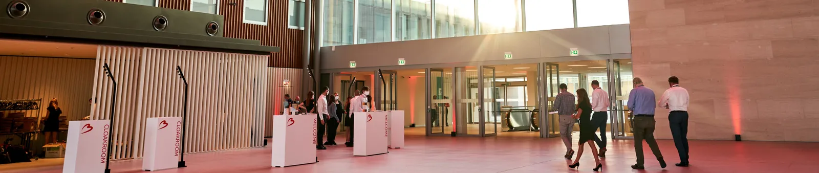 A modern building lobby with large windows and a pink floor. People are checking in at counters marked "Cloakroom" and "Welcome."