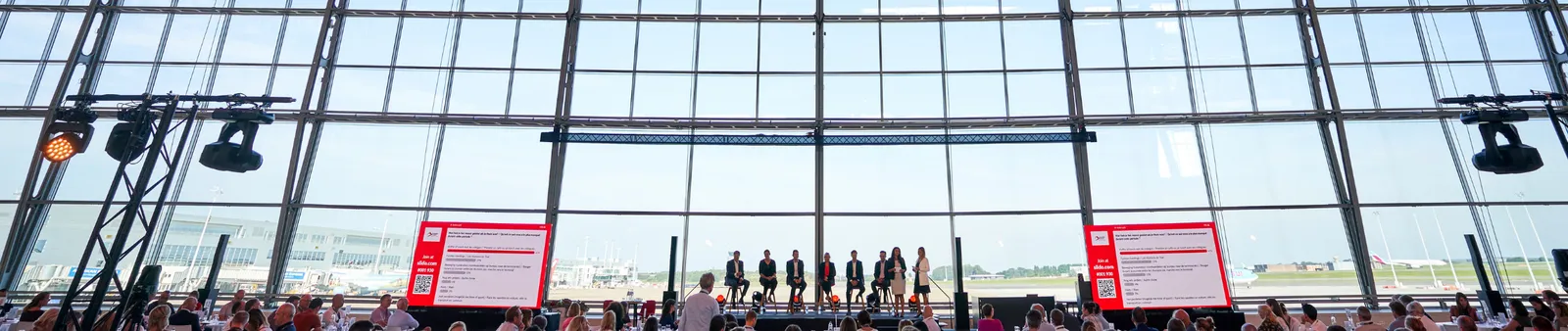 A conference in a large, glass-walled venue with speakers on stage and attendees seated at round tables, facing large display screens.