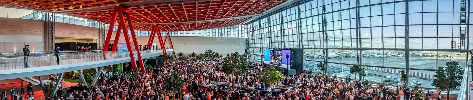 A large crowd gathers in a brightly lit airport terminal with tall glass windows overlooking the tarmac, featuring red architectural beams and greenery.