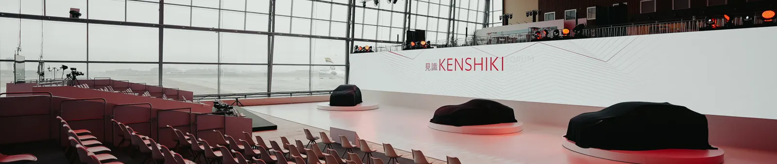 An empty presentation hall with a large screen displaying "Kenshiki." Three covered objects on podiums are in the center, surrounded by seating.