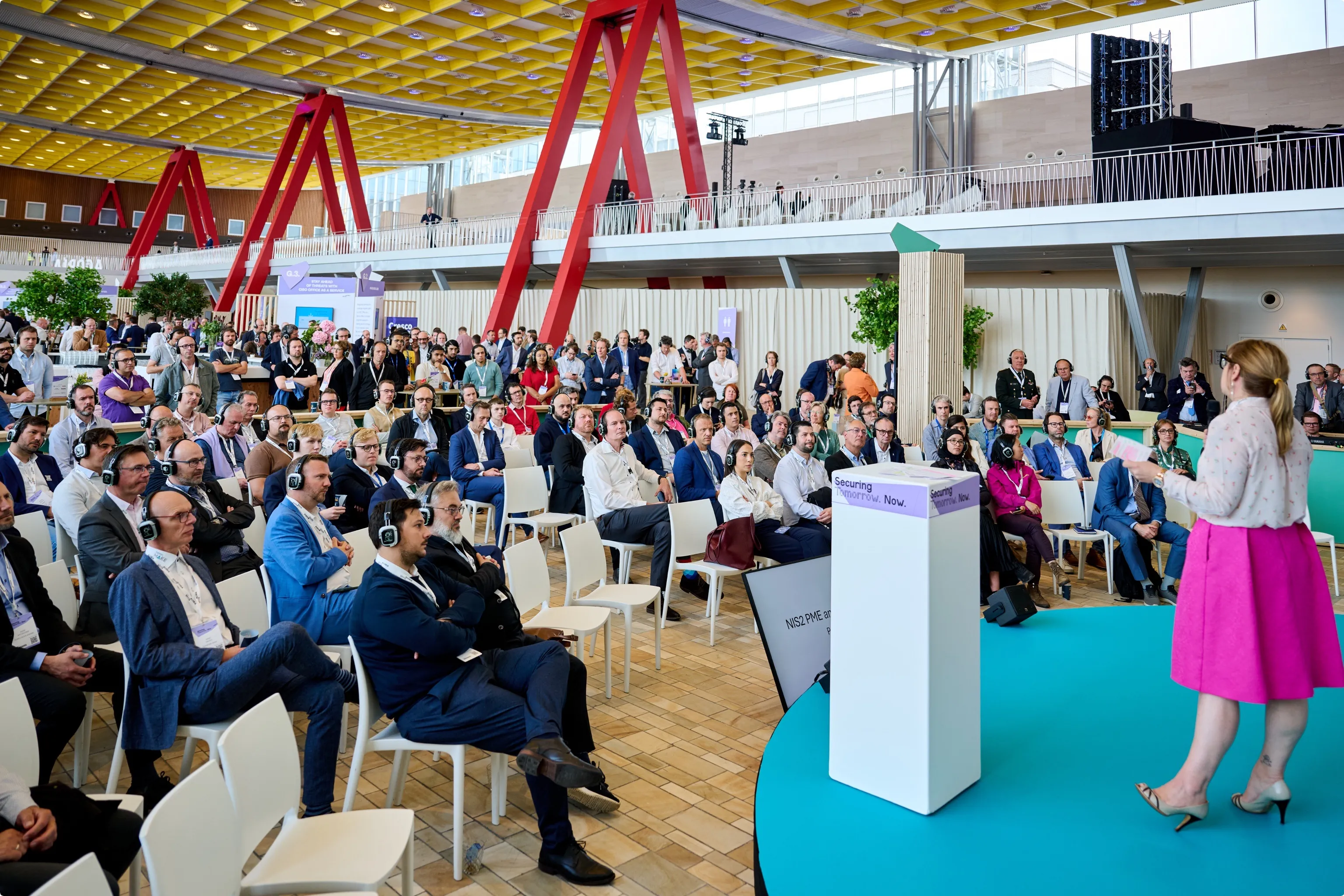 A woman speaks on stage to a seated audience wearing headphones, in a conference setting with a modern design and bright colors.