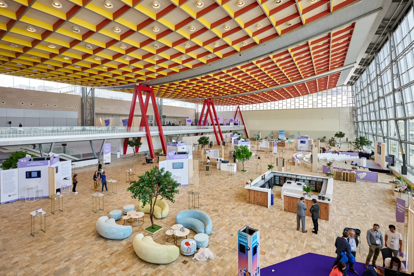 Spacious convention hall with modern decor, colorful ceiling, several booths, seating areas, and people networking in a bright and airy environment.