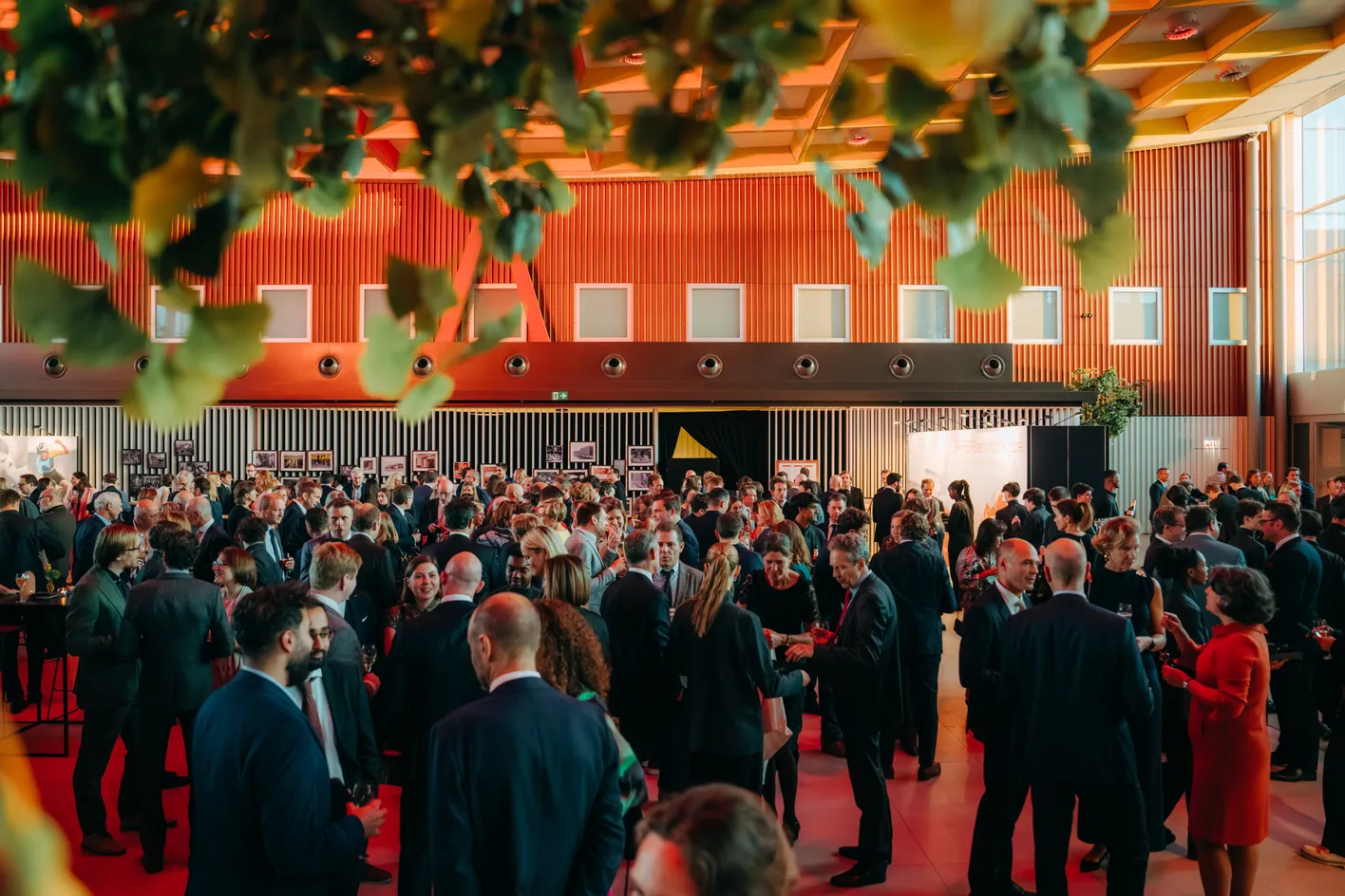 A large group of people in formal attire mingling at an indoor event with red lighting and greenery overhead.