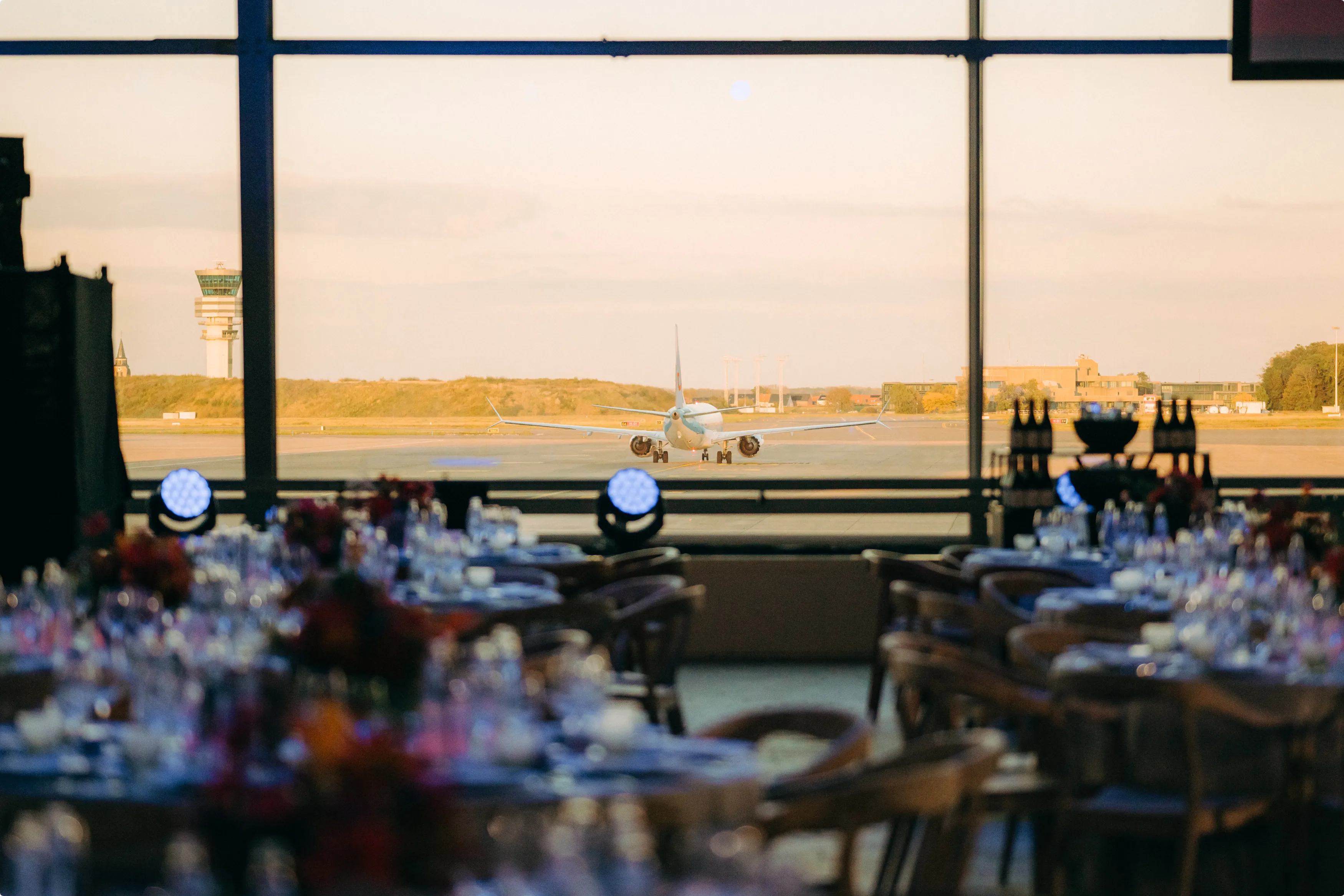 An airplane is visible through a large window at sunset, with elegantly set dining tables in the foreground.