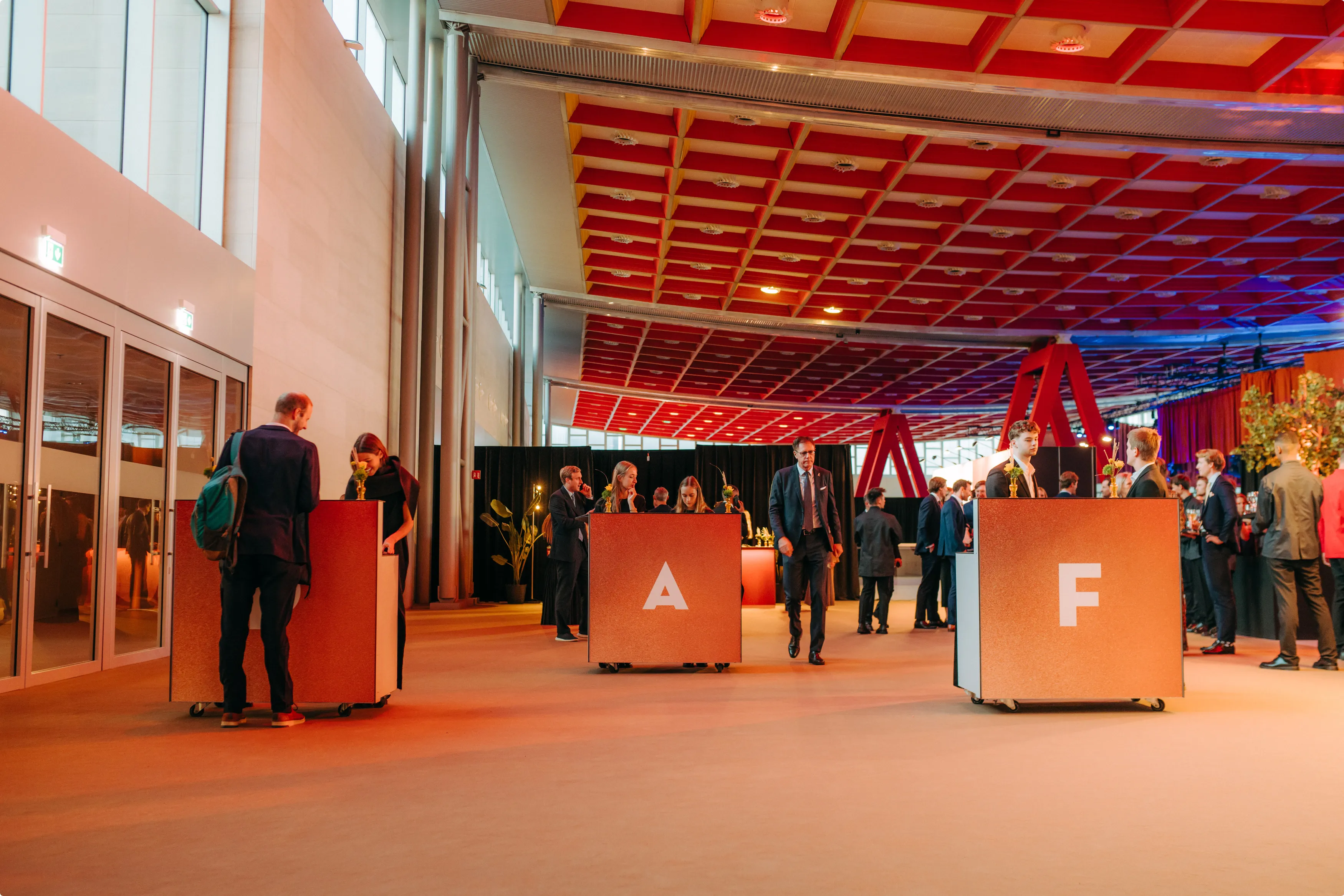 People in formal attire gather around large lettered boxes in a spacious, orange and red-lit room with a patterned ceiling and large windows.