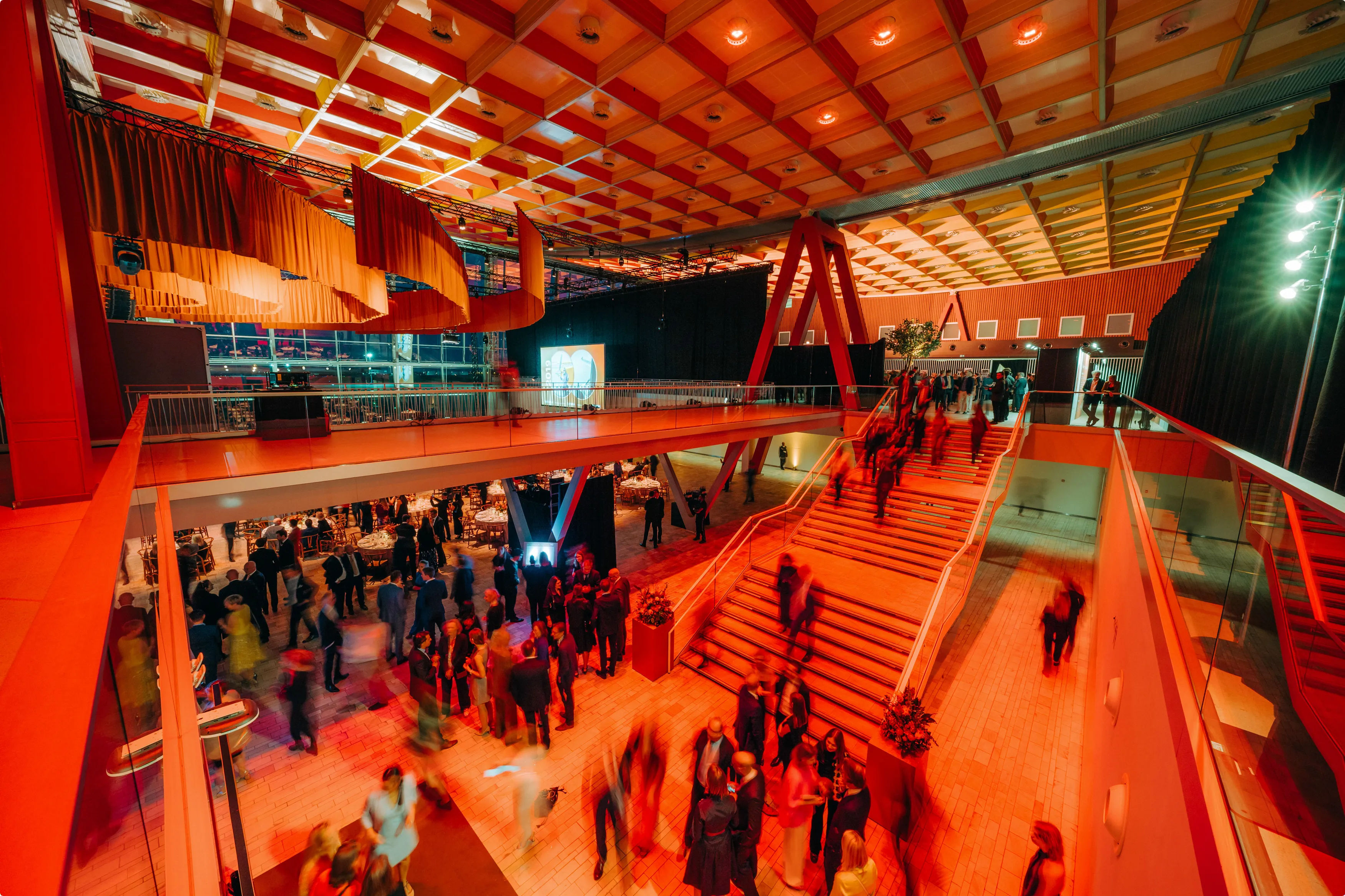 A vibrant indoor event space with people socializing, illuminated by warm red lighting, featuring a staircase, high ceiling, and a partially partitioned area.