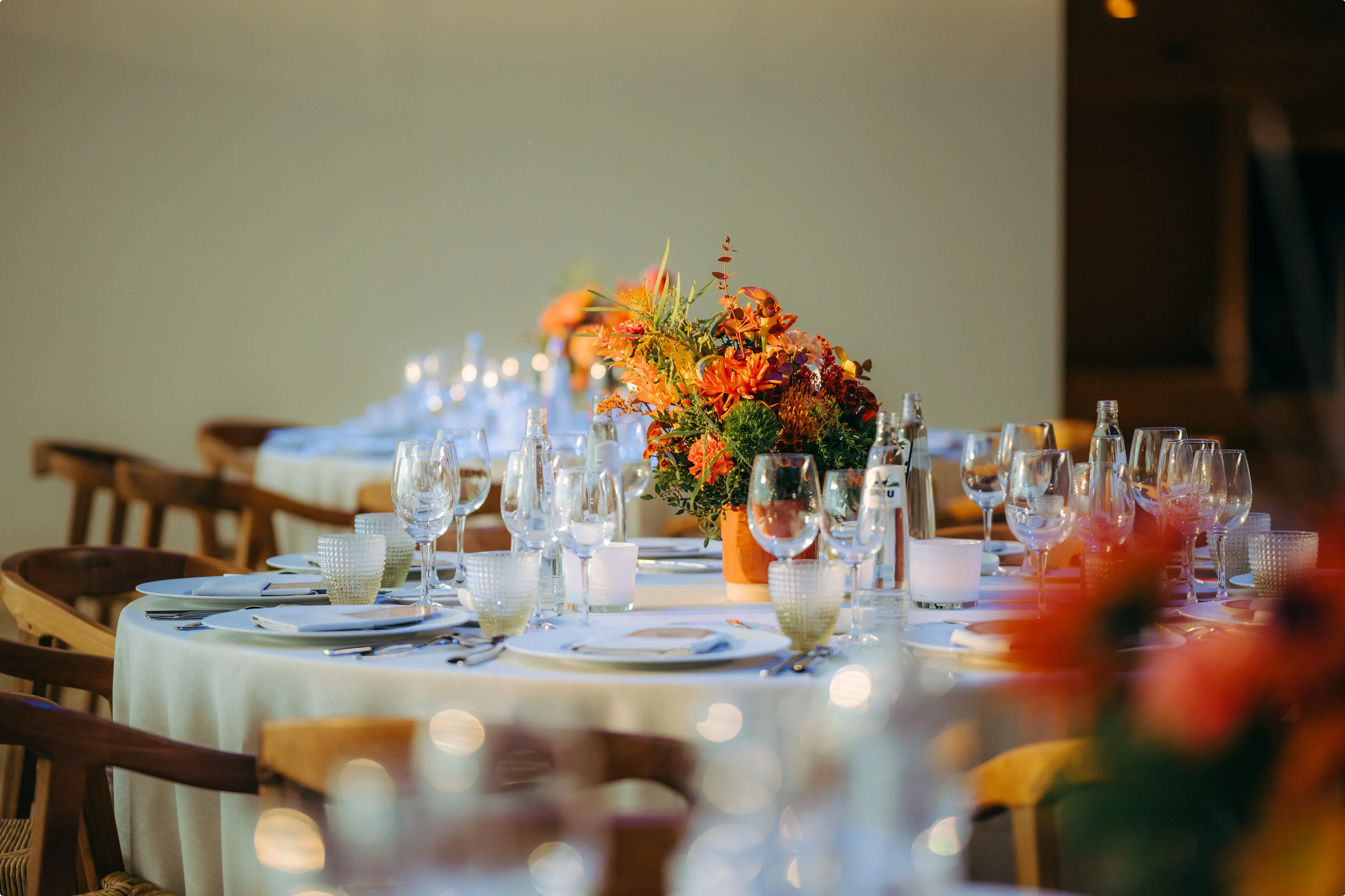 Elegant dining table with orange floral centerpieces, white tablecloth, arranged glassware, and cutlery set for an event or celebration.