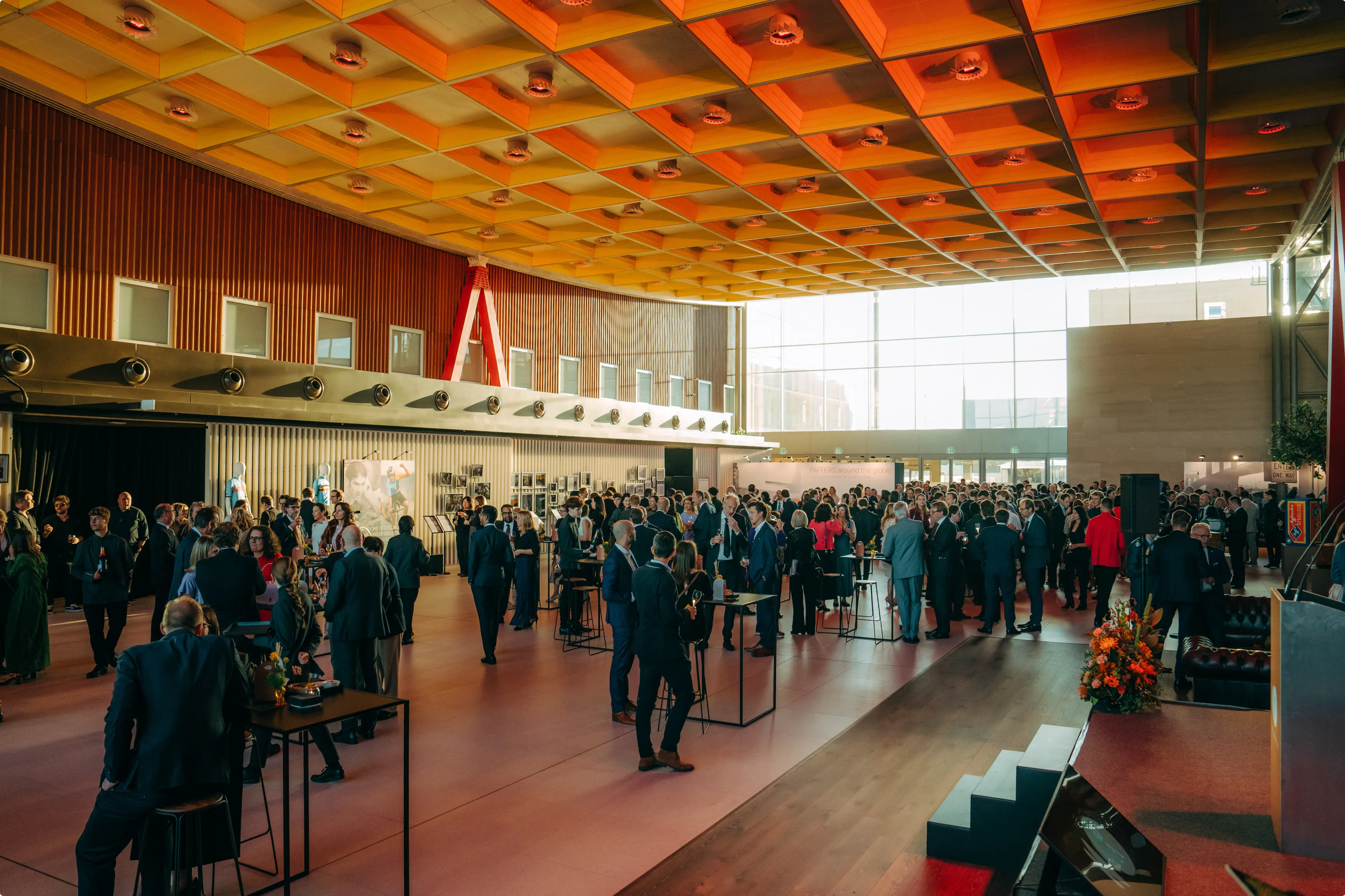 A large indoor event space with people mingling in business attire, illuminated by a warm, orange ceiling grid, with windows letting in natural light.