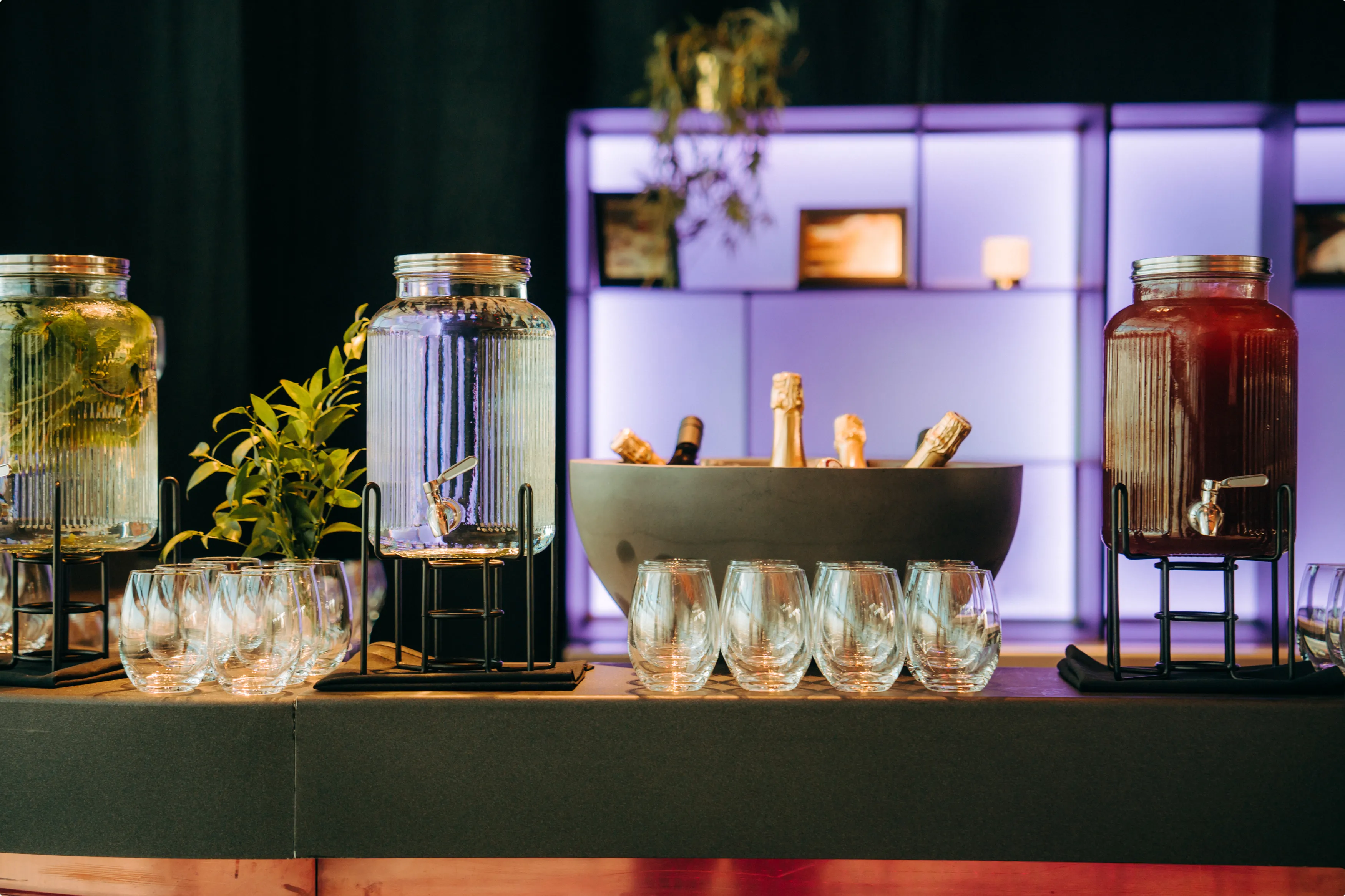 Glass dispensers with drinks, empty glasses, ice bucket with bottles, and plants on a table with a purple-lit shelf backdrop.