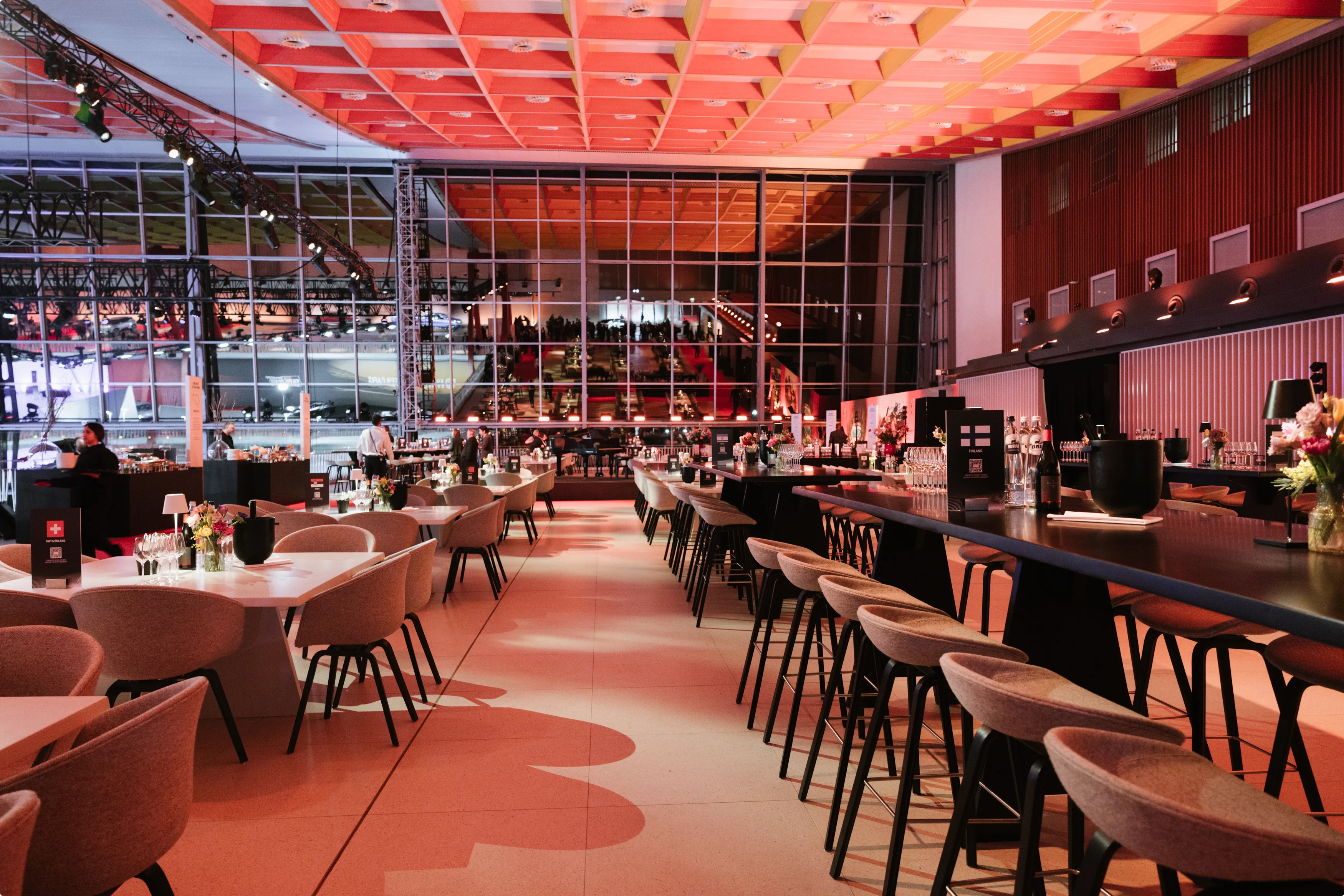 Spacious modern venue with stylish seating, tables, and a long bar, featuring large windows, warm lighting, and decorative flowers.