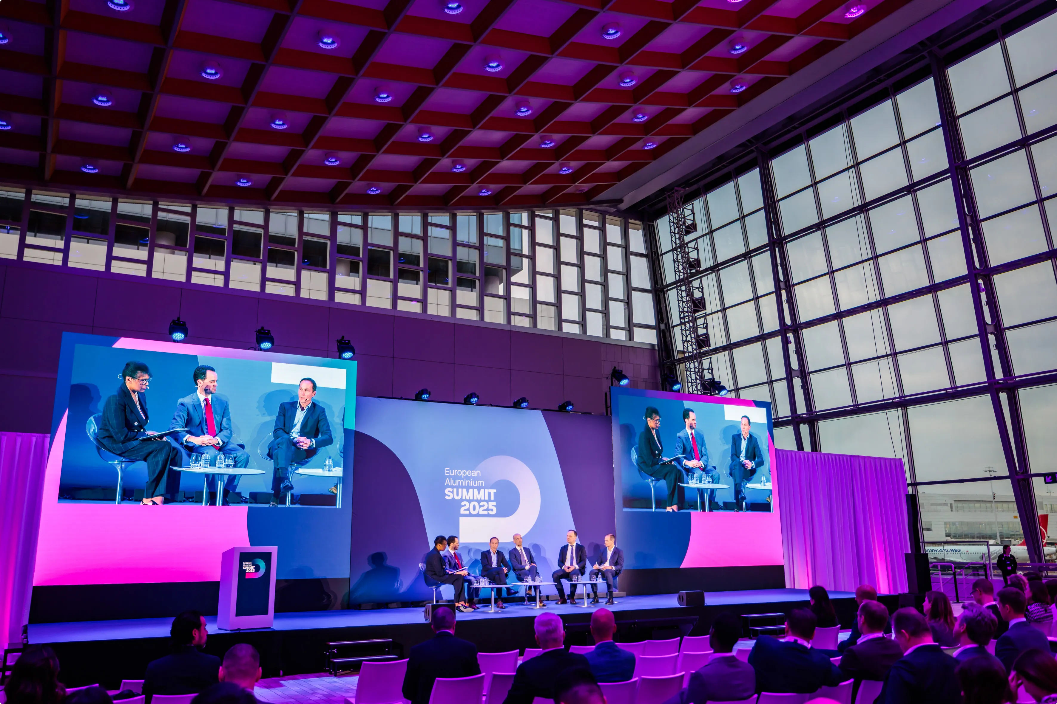Conference room with a panel discussion at the European Aluminium Summit 2025. Large screens display panelists against a backdrop of large windows.
