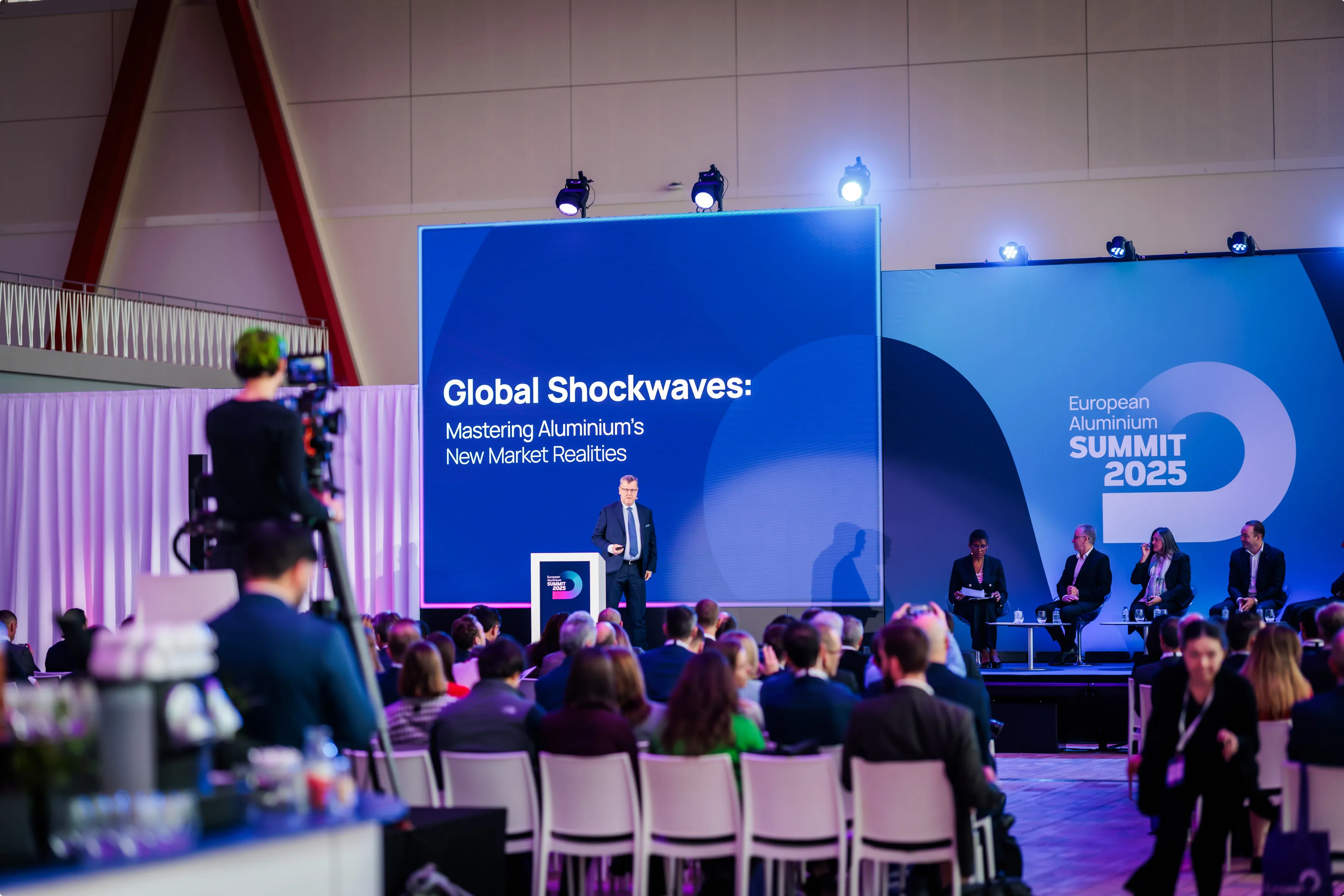 A speaker presents at the European Aluminium Summit 2025, with the topic "Global Shockwaves" displayed on a large screen in front of an audience.