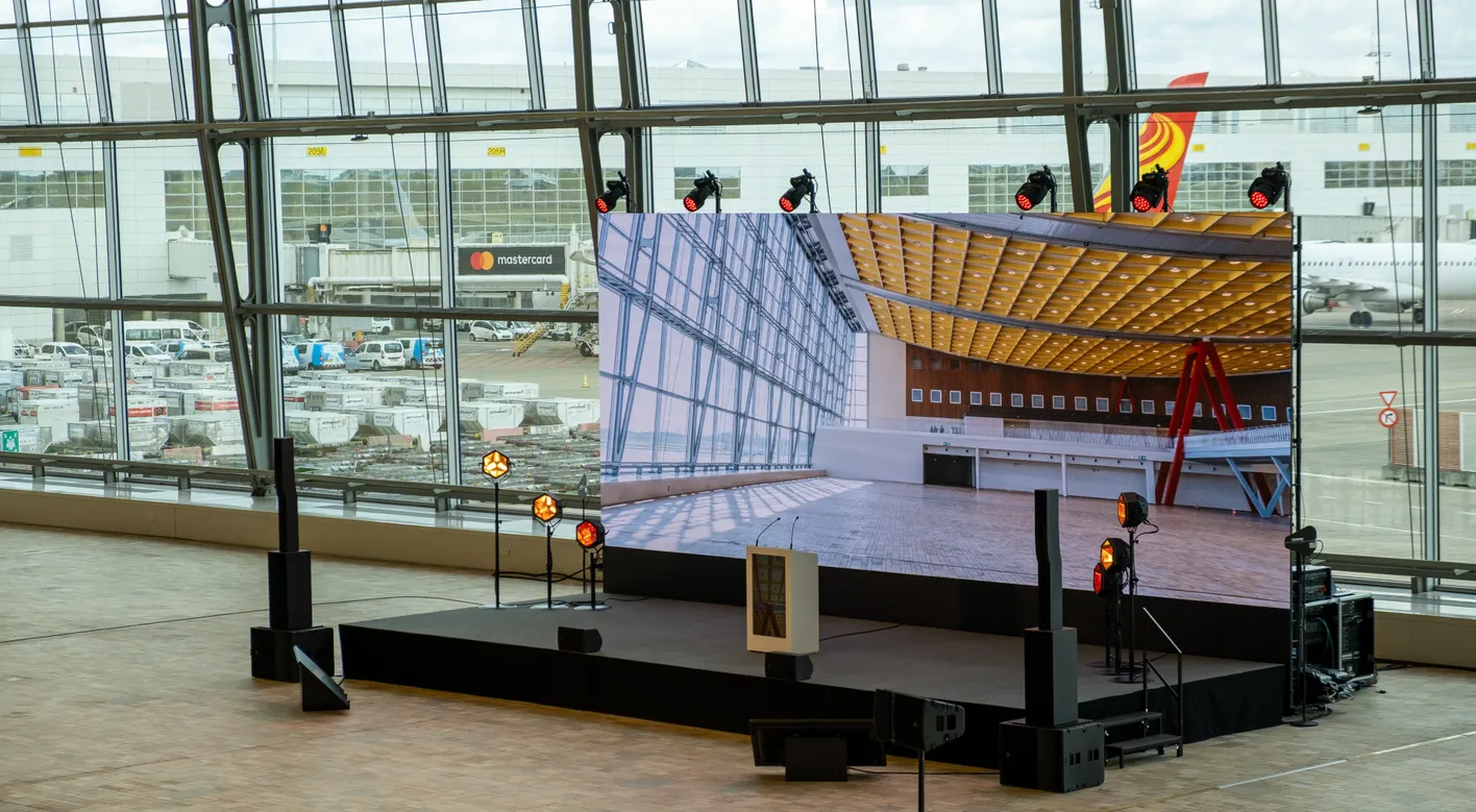 An airport terminal with a large screen displaying an interior image, surrounded by stage lights. Vehicles and planes are visible through windows.