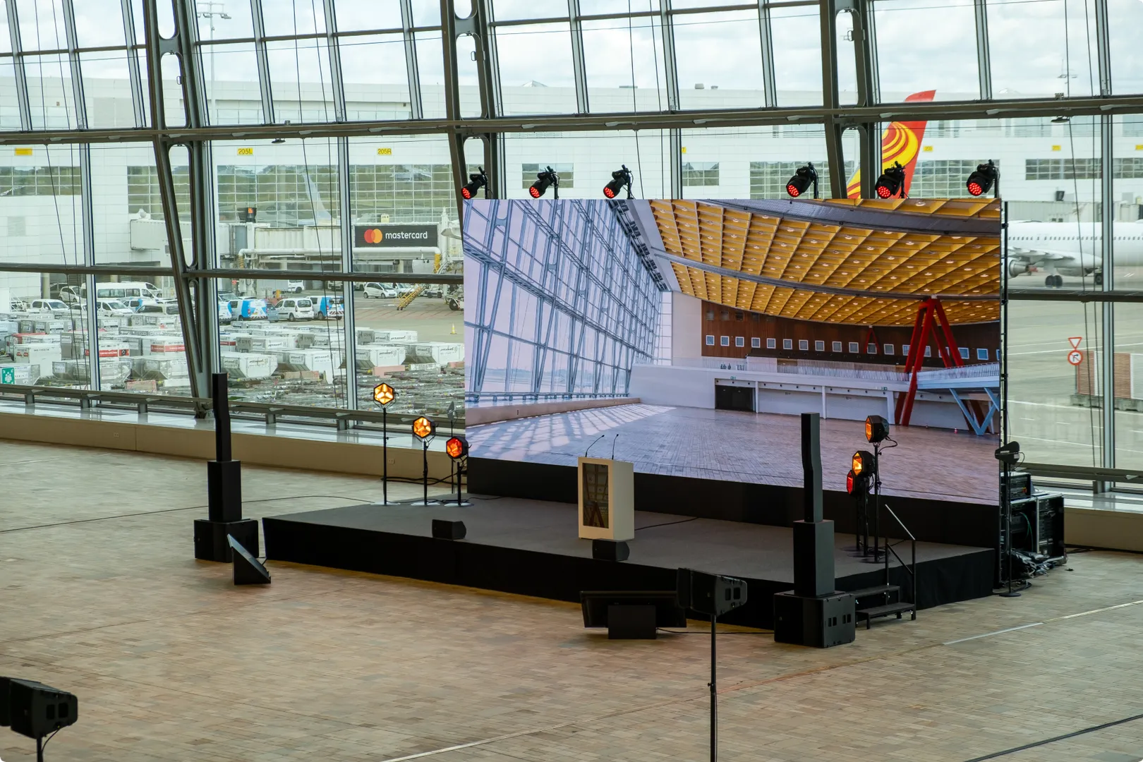 A large screen on a stage inside an airport terminal displays an image of an industrial interior. Airfield and parked cars visible through windows.