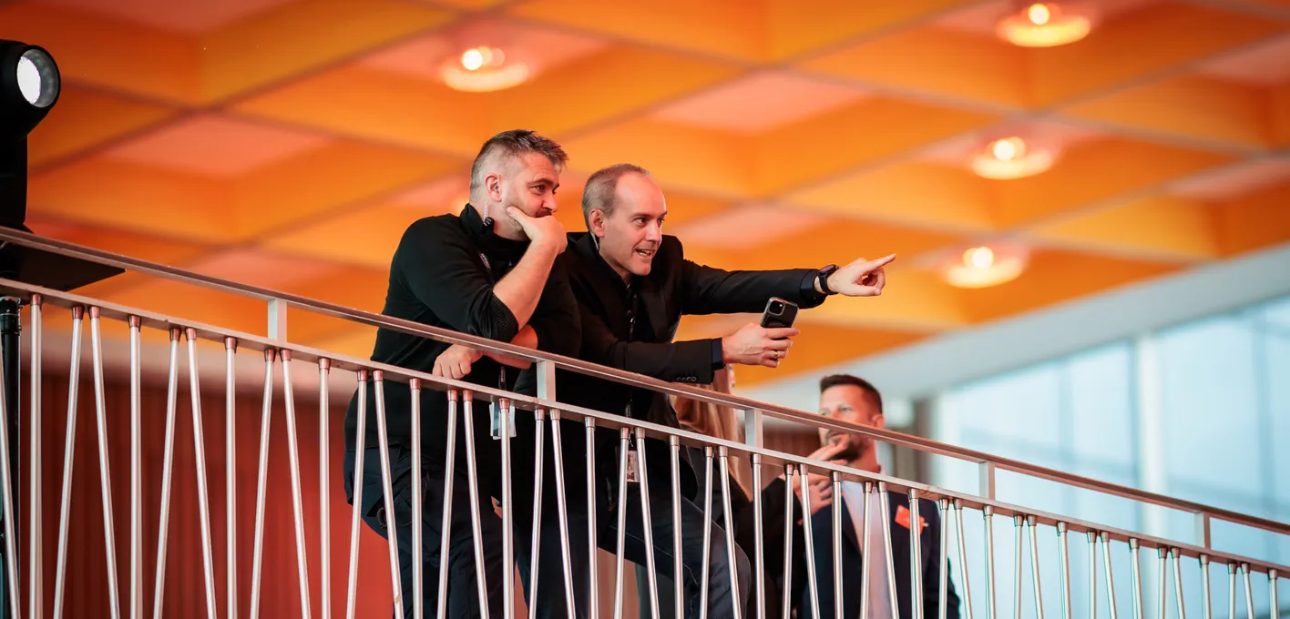 Three men stand on a balcony, one pointing with excitement while holding a smartphone, under a warm, orange-lit ceiling.
