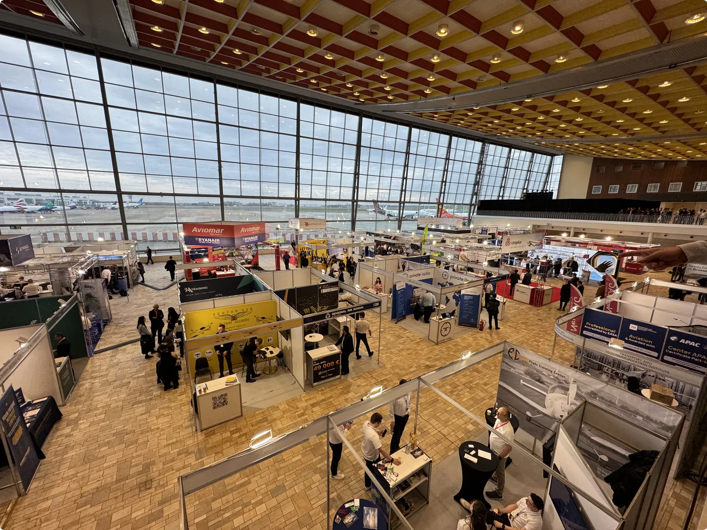 A busy exhibition hall with numerous booths and attendees, set inside an airport terminal with large windows overlooking parked airplanes outside.