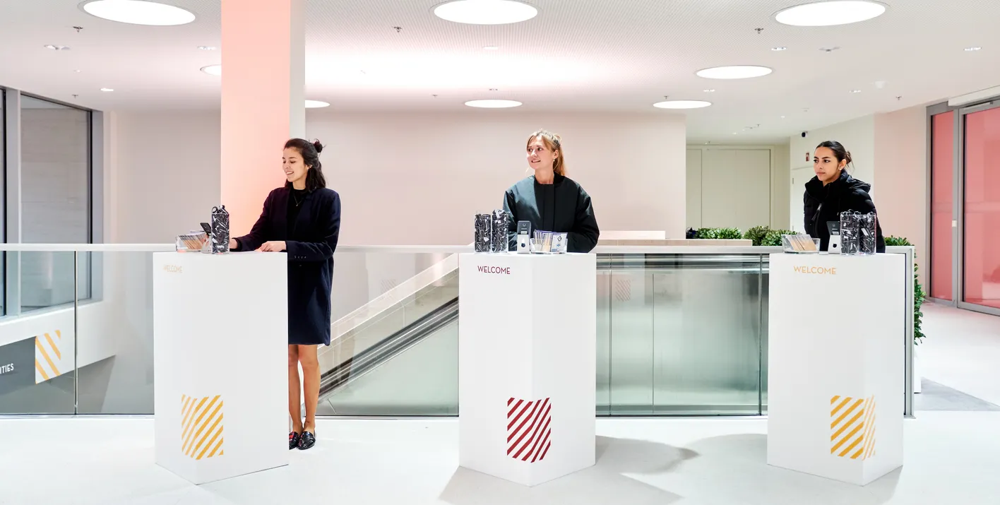 Three women stand behind separate welcome desks in a modern, brightly lit interior.