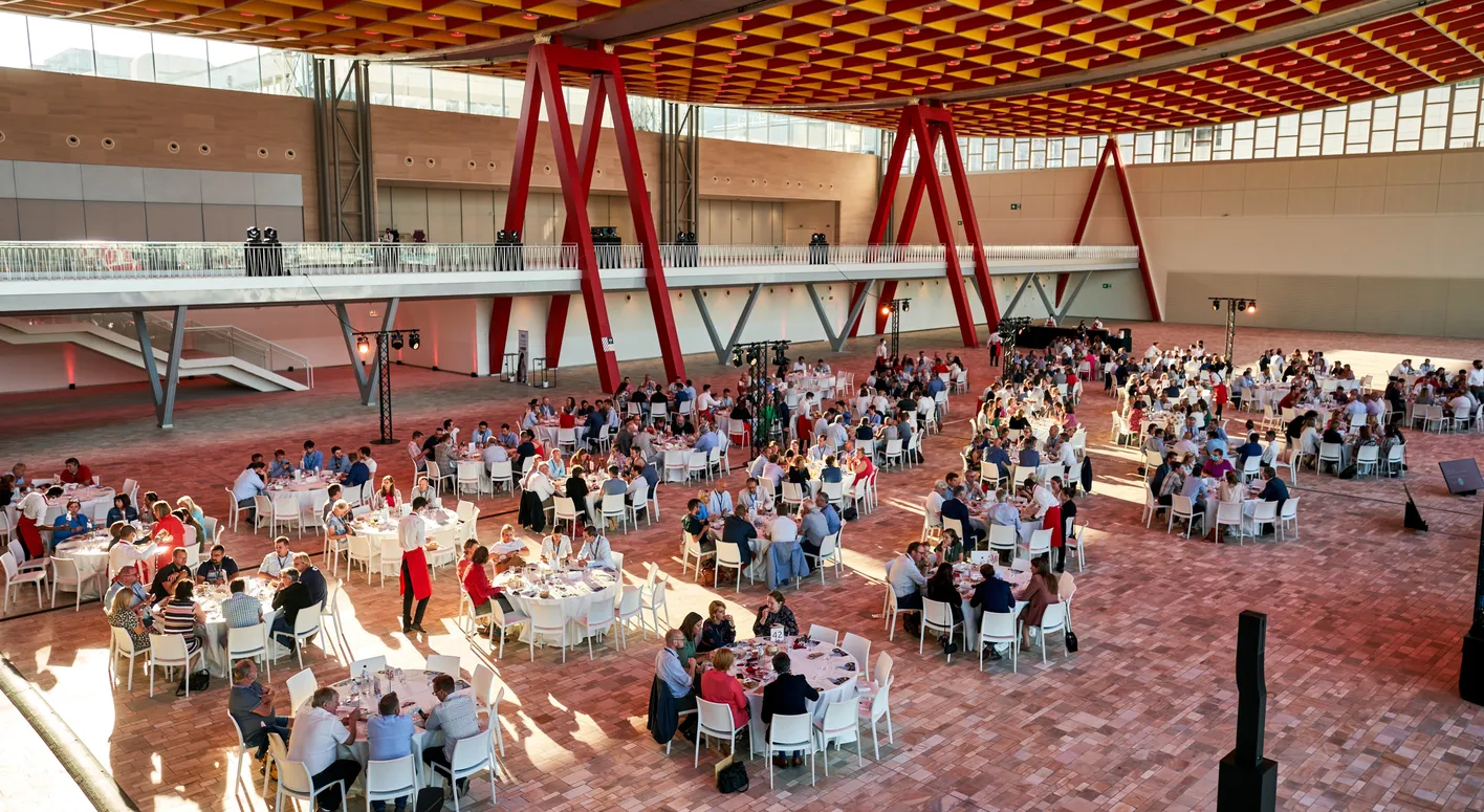 Large indoor event with numerous round tables and seated guests under a red and yellow ceiling. The room has a spacious and modern design.