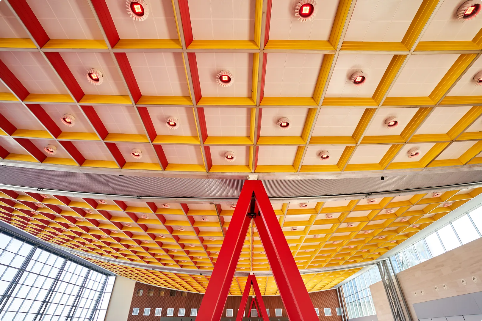 A colorful ceiling with a red and yellow grid pattern supported by red triangular beams, in a spacious room with large windows.