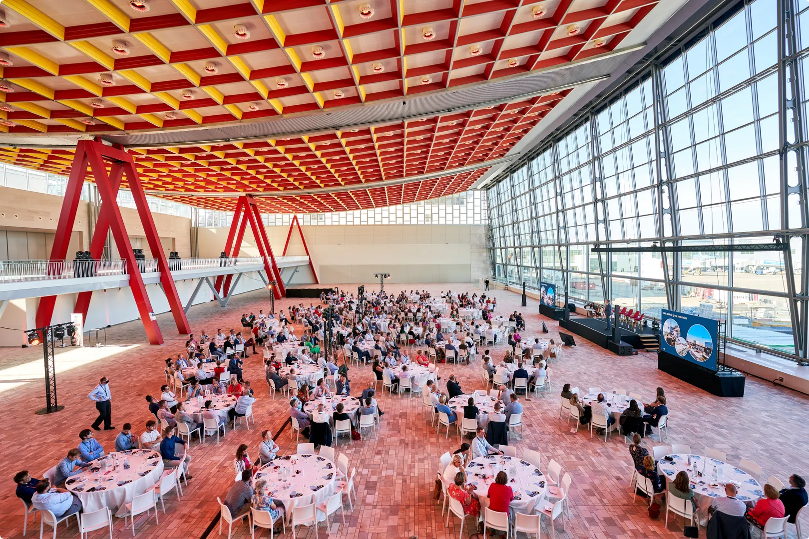 A large conference hall with a colorful ceiling, filled with round tables and attendees, and a stage with screens near a wall of windows.