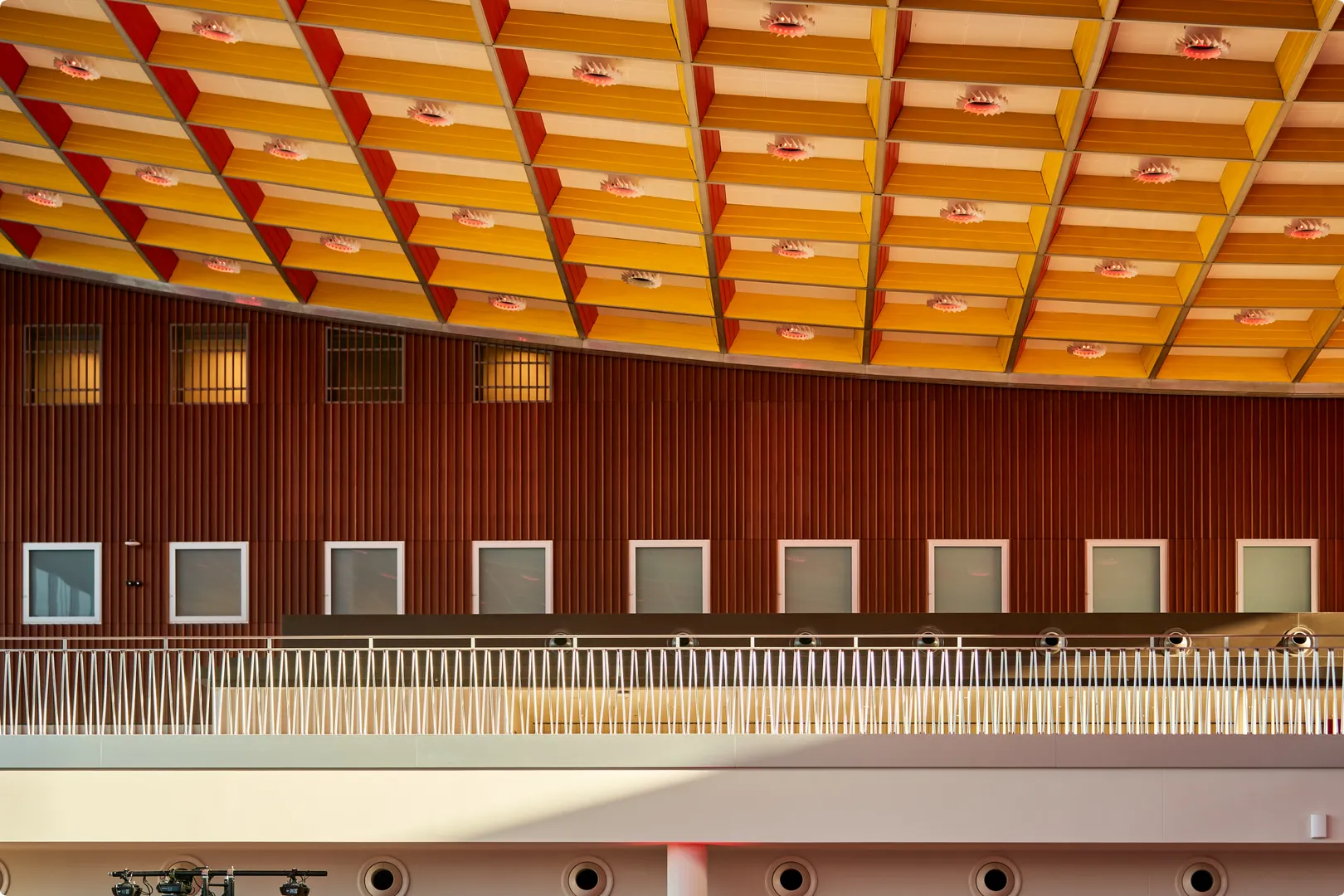Interior of a building with a geometric, colorful ceiling and a wooden-paneled wall featuring multiple square windows and a white railing.